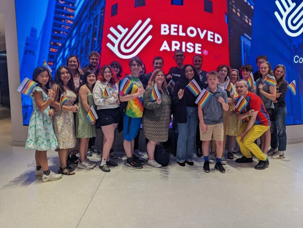 3201 | GLAAD A group of about 30 youth and adults holding rainbow flags, standing in front of the Beloved Arise logo at the exterior of the NASDAQ building.