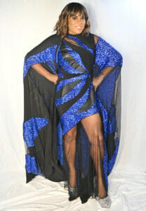 Tona Brown image | GLAAD Tona Brown, Black skin, with light brown hair, smiles a toothy smile under her bangs. She is wearing a shorter blue and black dress that drapes over her shoulders and arms like a cape. She stands in front of a white back drop with her hands on her hips. She stands proud.