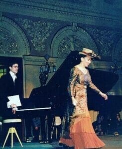 Performance in Prague | GLAAD Nina Kennedy, in a long warm-colored gown walks on stage in Prague to perform. The stage is dark and she wears a short top hat. Behind her is a man in a black composer's coat and a white shirt holding papers. There is a stool and instruments sat across the stage.