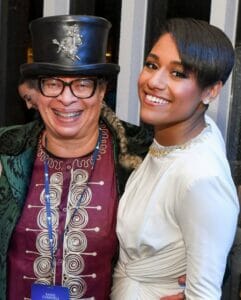 Nina Kennedy and Ariana Debose | GLAAD Nina Kennedy, light brown skin, with rectangular glasses smiles in a black top hat and black leather coat over a red shirt or dress, next to a smiling Ariana Debose, who is black with dark brown hair, wearing a white dress.