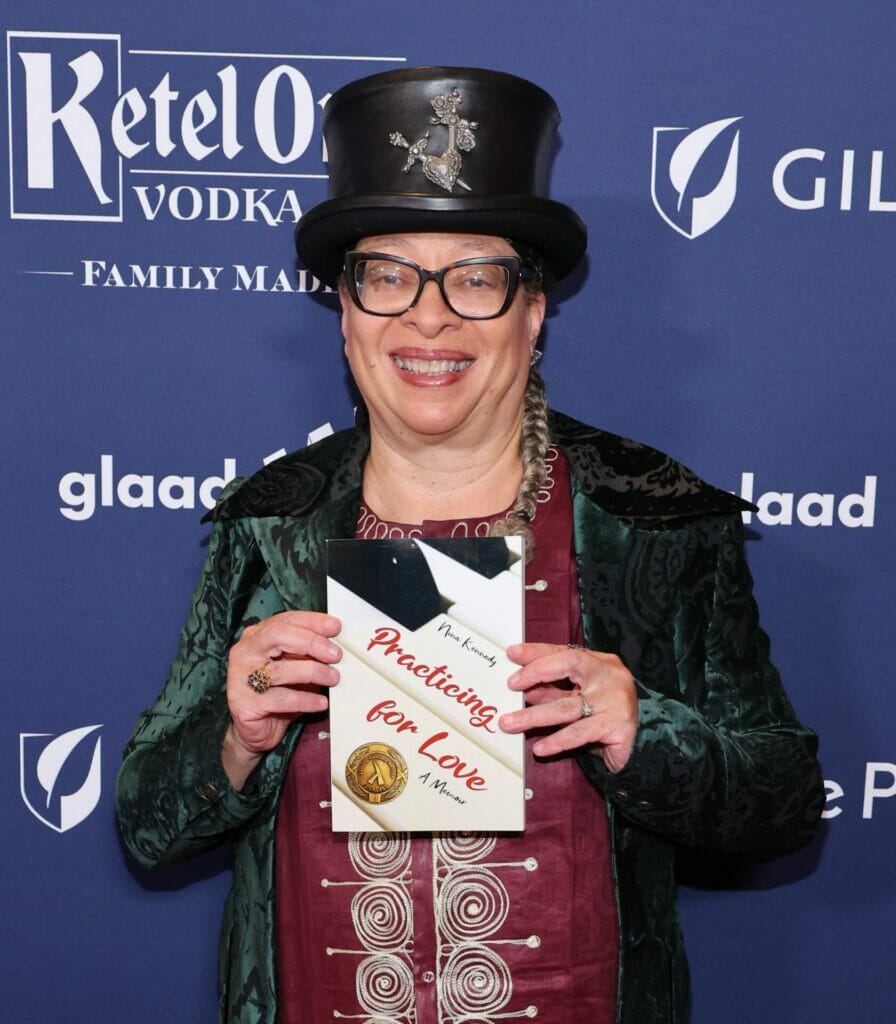 Nina Kennedy, in a black top hat, with light brown skin, wears a black leather (appearing) coat over a red shirt with a white design. She stands against a blue, GLAAD Media Awards backdrop holding part one of her memoir "Practicing for Love: A Memoir"