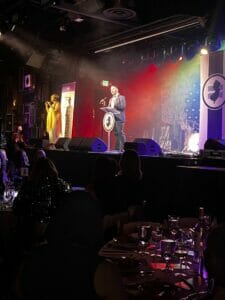 GSE Executive Director Christian Fuscarino Equality Ball 2023 address | GLAAD Garden State Equality Executive Director Christian Fuscarino gives 2023 Equality Ball address in dark suit with a white button up. His hand lightly extends out as he speaks to the audience under rainbow lights. The silhouettes of the audience can hardly be seen.