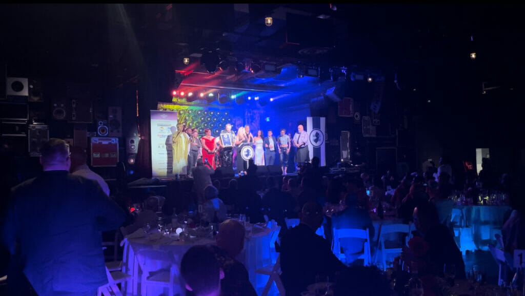 GLAAD Media Institute Alumni Visiting Nurse Association Community Health Center of Asbury Park stand in a row on stage for winning Garden State Equality's the Corporate Responsibility Award. The stage is lit in rainbow lights while the crowd is lit in blue and purple, while sitting at white round tables