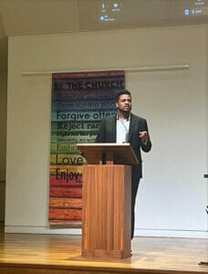 Rev. Redeem Robinson | GLAAD Rev. Redeem Robinson, Black, with short black hair, stands behind a honey brown wooden podium in front of a Pride banner.