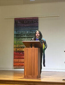 Bishop Allyson Abrams | GLAAD Allyson Abrams, Black, with greyish black hair, stands behind a honey brown wooden podium in a progressive pride sash in front of a Pride banner.