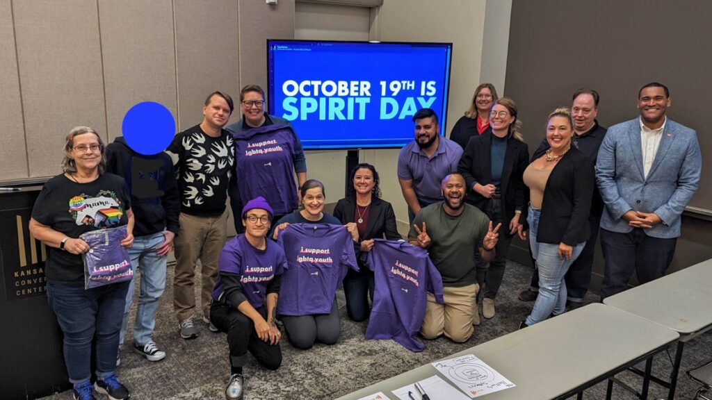 All participants of training come together for a picture with the GLAAD Media Institute (Lana, Ross and Jerrell). Many are wearing purple for Spirit Day, the world's largest anti-bullying campaign in support of LGBTQ youth. The room is grey and people surround a monitor which reads "SPIRIT DAY OCTOBER 19"