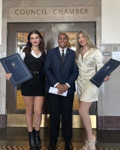 LGBTQ History Month | GLAAD Tristan Young (left), white with long brown hair smiles while wearing a black dress with a long sleeve white shirt under neath holding the historic resolution that she is the second trans woman to be honored the title of homecoming queen, and she stands next to Justice Horn (center), black with short brown hair, wearing a blue suit with a white collared shirt underneath and a dark tie, stands with his hands folded and smiling wide. He stands too with Landon Patterson (right), white, long blond hair smiles wearing a white dress also holding the city resolution that states she is the first trans woman to be voted homecoming queen. They both have their arms around each other and stand in front of the Kansas City Counsel Chambers; photo courtesy of Justice Horn