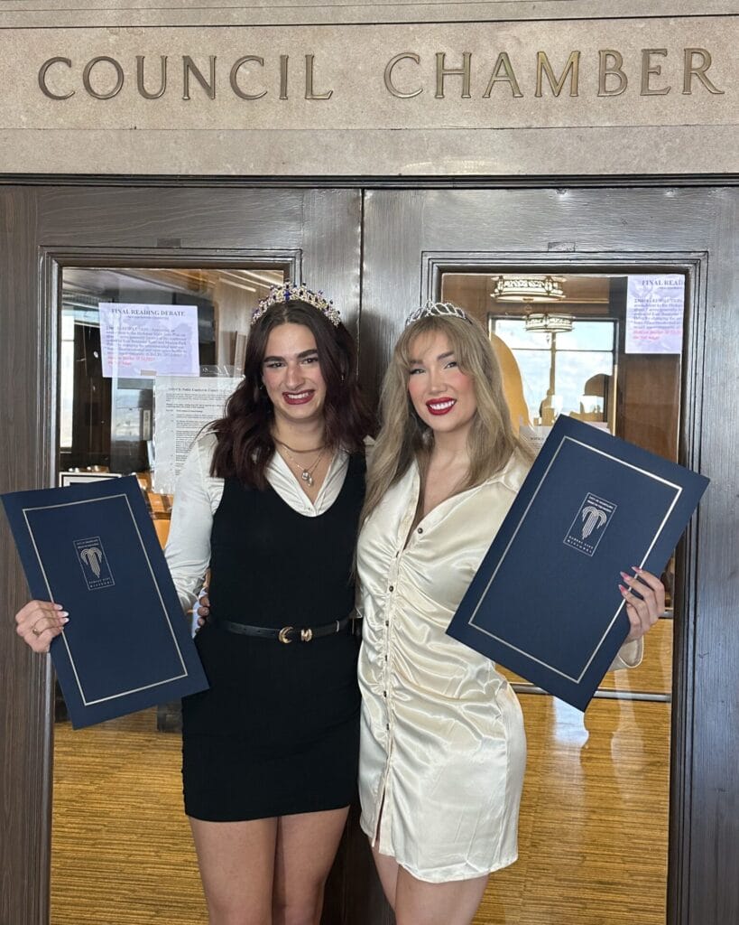 Tristan Young (left), white with long brown hair smiles while wearing a black dress with a long sleeve white shirt under neath holding the historic resolution that she is the second trans woman to be honored the title of homecoming queen, and she stands next to Landon Patterson (right), white, long blond hair smiles wearing a white dress also holding the city resolution that states she is the first trans woman to be voted homecoming queen. They both have their arms around each other and stand in front of the Kansas City Counsel Chambers; Photo courtesy of Justice Horn