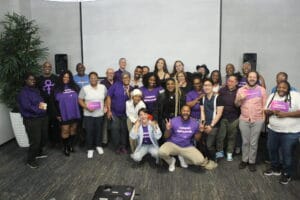 Las Vegas GMI Alum Group Photo | GLAAD Las Vegas LGBTQ advocates take a group photo with the GLAAD Media institute Education and Advocacy Team. Almost everyone is wearing purple and is smiling with arms around one another. About half of people in the photo are wearing purple in the name of Spirit Day. The background and carpet are grey. The wall light grey and the carpet dark.