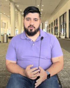 Screenshot 2023-10-18 at 9.25.05 AM | GLAAD Alvaro Ontiveros Aguilar, tan complexion, brown hair and beard,and dark brown eyes, purple shirt with his hands on his lap. He is wearing dark blue jeans. The Kansas City Convention Center is in the background.