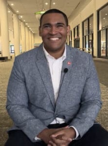 Screenshot 2023-10-18 at 9.25.39 AM | GLAAD Justice Horn, dark complexion, smiling with a bright smile, in a grey suit with a white shirt, hands folded on his lap, with a "KC" lapel pin on his suit. The Kansas City Convention hall is in the background.