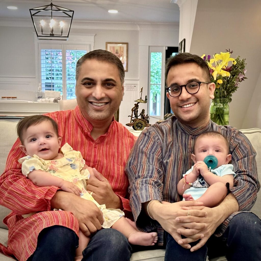 Parag Mehta (left), short black and grey hair, darker skin, in a red shirt button-up, smiling while holding his baby dressed in yellow with husband Vaibhav Jain (right), with black short hair, smiling with glasses, in a checkered black and brown shirt, holding their other child dressed in blue. Their home is white with yellow flowers in the background.