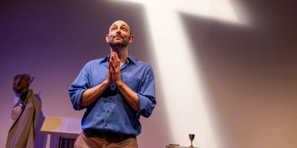 Matthew LaBanca poses with prayer hands in front of a projected image of a cross.