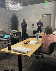 Screenshot 2023-10-04 at 2.10.47 PM | GLAAD André Wade, black, pruple and black shirt speaking into a microphone to the audience while standing next to Ross Murray, white, purple button down with black sports coat, in grey room with long light wooden tables.