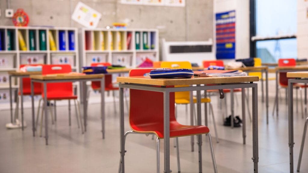 Colorful classroom with tables and chairs strewn throughout the classroom.The red seats are empty. The back of the classroom has rainbows above the desks with windows over to the left wall.