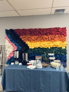 Shepherds Table Refreshments | GLAAD A paper mache progressive pride flag on the wall compliments hot water and coffee, cups and plates for food on a table with a blue table cloth.