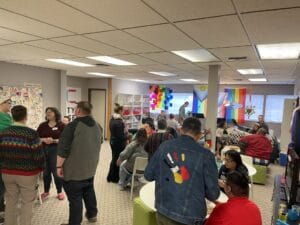 IMG_0894 | GLAAD A room full of people standing scattered around the new ministry. Pride flags surround the guests as well as food, table and chairs on a Sunday. The ceiling is white with panel LED lights. There are big windows with LGBTQ pride flags in the distant.