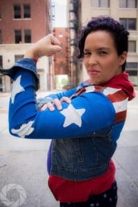 Fern 14 | GLAAD Fern Galindo, brown skin, short curly brown hair, wearing a red lipstick, and a red, white, and blue leotard mimicking the American flag. Fern has his arm curled in a flex