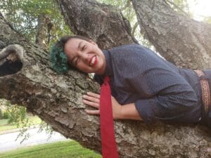 Fern 15 | GLAAD Fern Galindo, brown skin, green and brown curly hair, red lipstick, hugging a tree horizontally wearing a charcoal button up, a red tie dangles over their left hand.