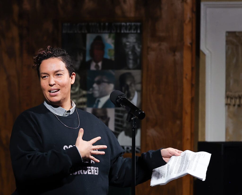 Fern Galindo, they/he, 38, brown skin, short dark brown, curly hair, wearing a dark sweater over a collared shirt is reading from a sheet of paper. They are looking over their right shoulder speaking with their hand on their chest. Behind them is a collage of peoples' pictures.