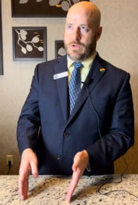 Casey Willits | GLAAD Casey Willits, white, bald, with a short brown beard, in a dark blue suit, a light green shirt, blue-speckled tie, with a Gainesville Pride pin, and stands over a beige, and black speckled granite countertop.