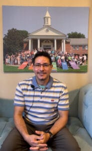 image_123650291 2 | GLAAD Saer Olavarria has brown, olive skin, with short dark hair. He wears rectangular shaped glasses, and has a mustache with a soul patch. He wears a blue and white striped polo shirt, with grey pants. He sits on a blue couch in Glendale United Methodist Church with a frame of members of the church behind him.