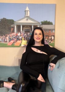 image_123650291 | GLAAD Ravanna-Michelle Menendez, she/her, has pale skin, she wears a skin tight, black top, with a score in the chest area just before the collar of the shirt. She has dark brown/black hair that flows past her shoulders, in a leather jegging with a thick black heal. She sits on a blue couch, behind her is a picture in front of Glendale United Methodist Church in Nashville.