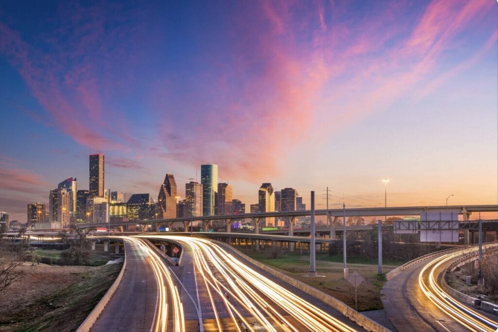 A blue and pink sky hang over the beautiful Houston skyline, with buildings in the distance and racing cars on the roads before the city streaming with white car lights