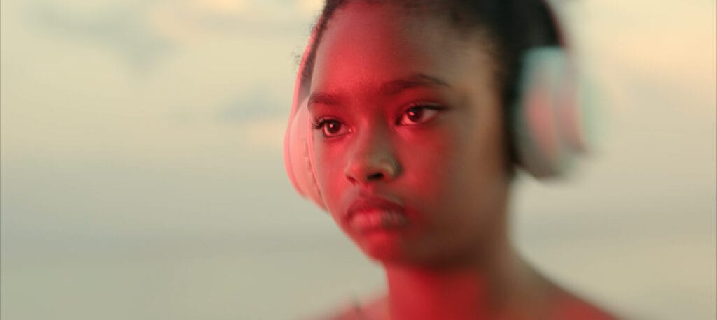 Young girl with neutral expression wearing large headphones in red light in front of cloudy sky.