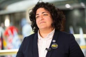 KP1_4701 | GLAAD Councilperson Liliana Bakhtiari, light brown complexion, bright eyes, with curly brown hair, stands in a dark suit with a white button up shirt with pins that say "Pride" on it. The background is blurred out.