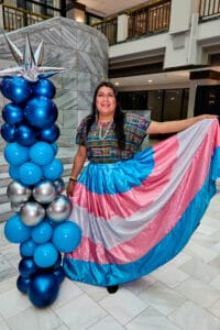 IMG_2830 | GLAAD Li Ann Sanchez, brown skin with long straight brown hair, stands next to a shiny, multi-shade blue, and silver balloon sculpture on the white marble floors of Atlanta City Hall with a trans pride flag skirt which she holds up at 90 degrees