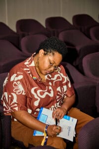 IMG_2722 | GLAAD Yemko Pryor, brown complexion, short brown hair with golden, circle frames, sits taking notes in the auditorium. They are wearing a brown button up shirt with white/cream floral-looking designs with burgundy seats in the background