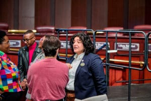 IMG_3088 | GLAAD Yemko Pryor, and Councilperson Liliana Bakhtiari stand around people in the City of Atlanta Council Chambers. Yemko is Black with short brown hair, wearing round glasses. They are wearing a colorful multi-colored pattern shortsleeve button up, and look right across from Bakhtiari who is speaking to a person in a red tshirt tucked into light blue jeans. Bakhtiari has fair eyes, and light brown complexion, with clurly medium length curly brown hair. They are wearing a blue sports coat over a button up.