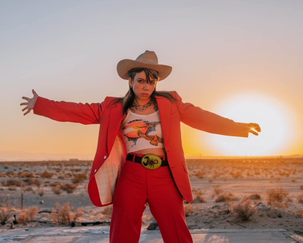 Alynda Segarra from Hurray for the Riff Raff stands with their arms out in front of a sunset wearing a cowboy hat and a pink suit