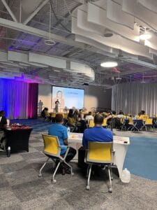 IMG_3558 1 | GLAAD The Center in St. Petersburg Florida. A room with bright overhead lights, grey walls and floor with a rainbow lit curtain just before the stage where people are presenting and discussing the Pride Amplified event. Yellow chairs and up to 40 people fill the room.