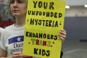 IMG_9915 | GLAAD Person, white with platinum, curly hair, shoulder length, wears a shirt, that says "Son't Mess with Trans Kids" and holds up a bright yellow 11x8 poster that reads "Your Unfounded Hysteria Endangers Trans Kids" in the Clifton Middle School gymnasium