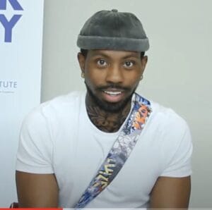 Screenshot 2024-06-16 at 32825 PM | GLAAD Charlie Xile, Black with a grey cap on his head. He has a dark brown beard, and smiles at the camera with bright brown eyes wearing a white t-shirt.