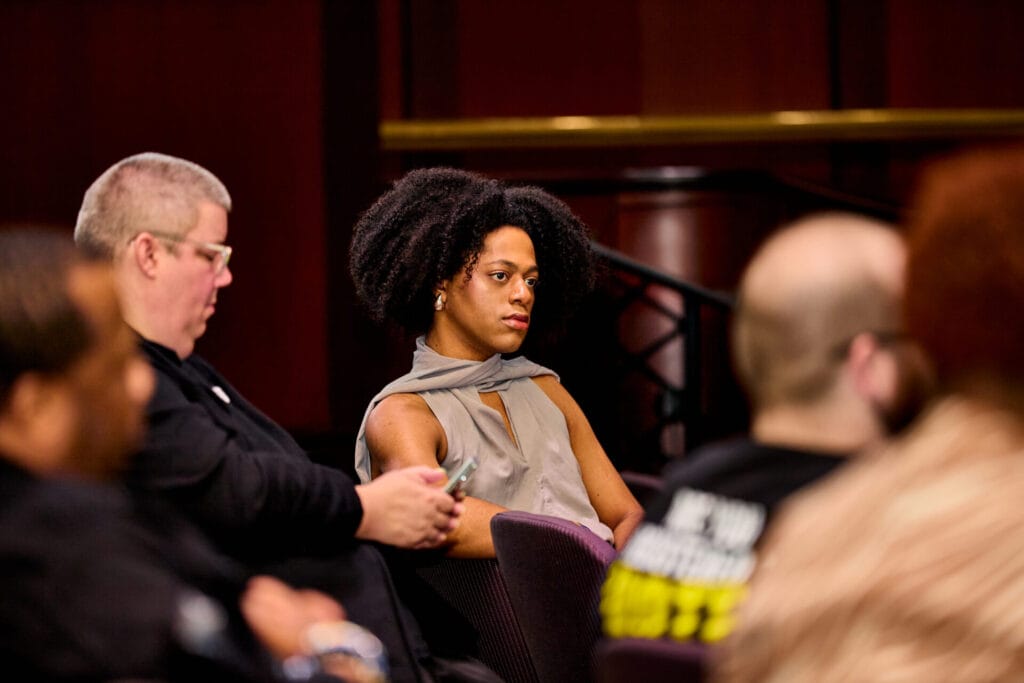 Tiana Tukes, Black woman with a longish, black afro sitting just above shoulder length, wears a grey garment. She sits in the Council Chambers with others as they pay attention to the speaker. The room is full of dark stained wood.