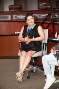 KP1_4674 | GLAAD Aubri Escalera, dark brown hair tied in a bun, light brown skin, wearing a v neck black dress with a elaborate blue necklace and sits in a white chair with her legs crossed in the Council Chambers