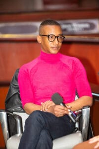 KP1_4792 | GLAAD Victor Jackson wearing a black leather jacket and pink shirt and blue jeans, he has short brown hair, and brown skin, and black rimmed glasses sits holding a mic listening to Park Cannon and Liliana Bakhtiari