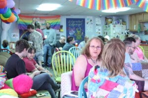 IMG_9511 | GLAAD Laurel Walker, white, wearing a pink dress, sits in a chair, her hair brown, and long, her eyes blue, and sits with a grand rapids community inside the Pride Center. Colors are everywhere, streamers, flags, signs, and posters, drawings by kids, and other artists. .