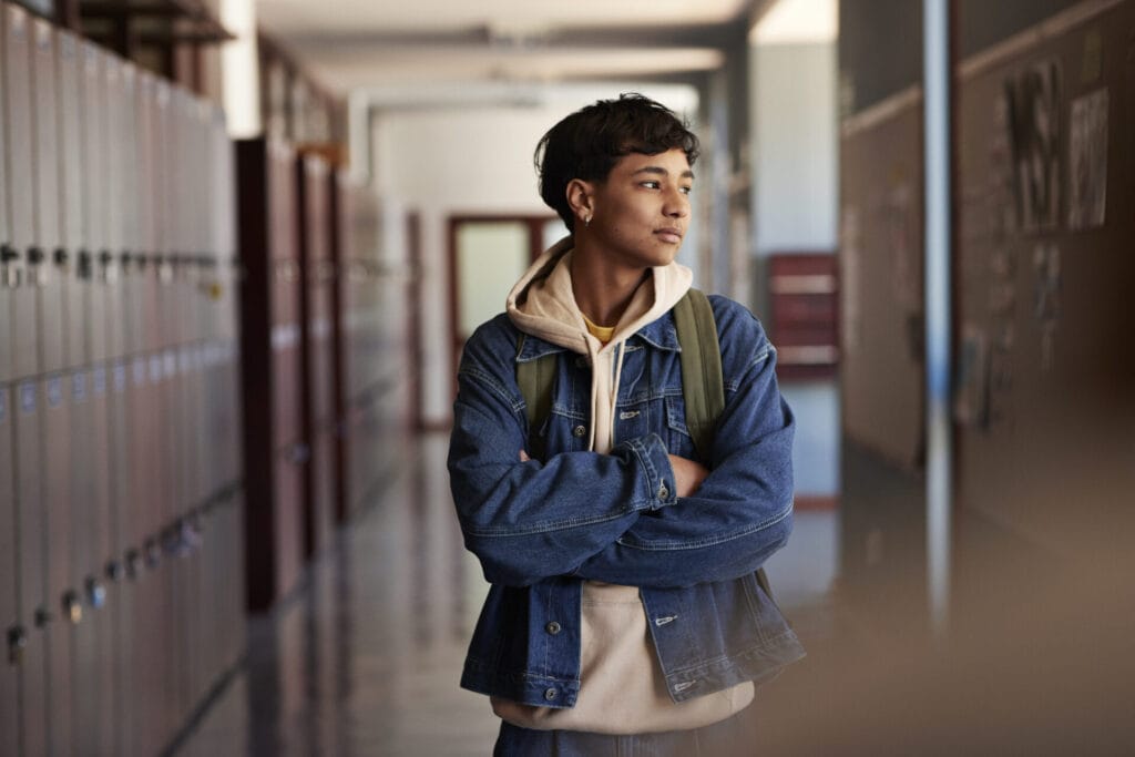 Teenage boy wearing denim jacket standing with arms crossed looking away in corridor at high school