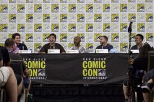 IMG_2943 | GLAAD Five transmasculine individuals sitting at a table on a stage. The backdrop and tablecloth feature repetitive blocks of San Diego Comic Con's logo.