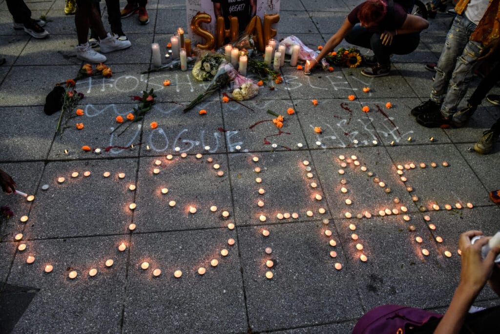 On the piers in downtown NYC, mourners gather to remember O'Shae Sibley. On the head of the pier people create a memorial for O'Shae. his name is spelled out in candle light, with feet circling, flowers in the distance ounder a photo of Mr. Sibley taken from us on July 29, 2023.