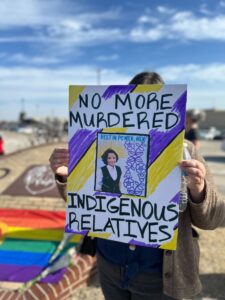 IMG_1837 | GLAAD "No More Murdered Indigenous Relatives" reads the sign the person is holding. It is in yellow, purple, white and black horizontal strips. Behind the person is Owasso High School, a blue sky with scattered clouds.