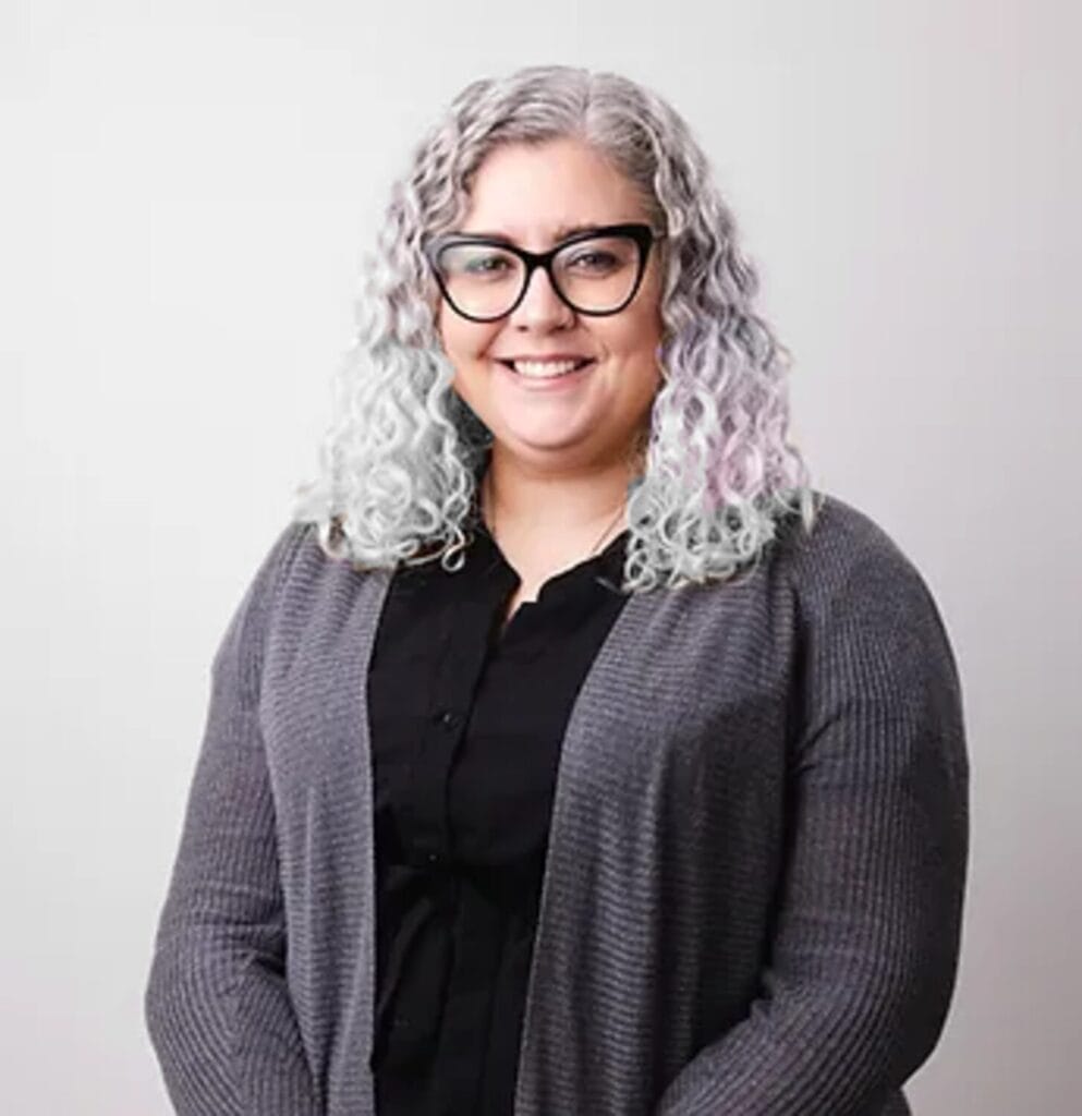JessickaHeadshot | GLAAD Jessicka Spearman, white, brown eyes, large black fram eglasses, shoulder length wavy gray hair, gray sweater and black shirt with off-white background