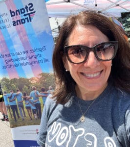 IMG_4415 | GLAAD Roz Keith, white, brown long hair with big, round brown sunglasses, stands smiling in front of a "Stand With Trans" banner