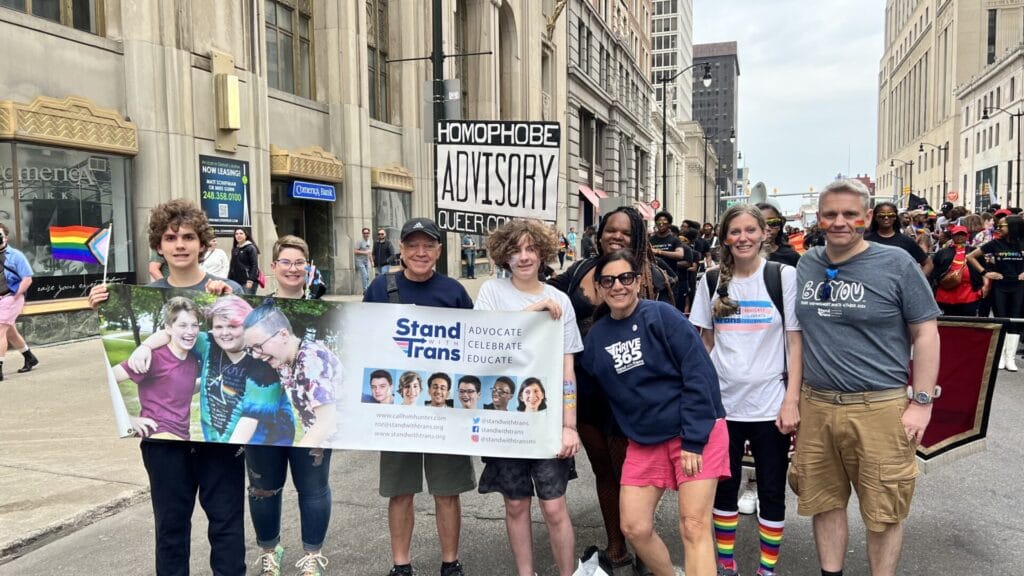 The group "Stand with Trans" at an action. About eight people stand in line smiling, holding a sign that reads: "Stand with Trans"
