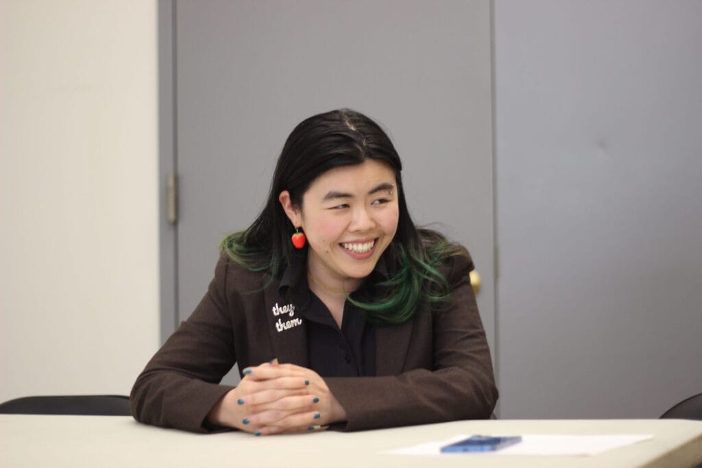 IMG_1242 | GLAAD Ginger Yifan Chen in a brown jacket with a "they/them" patch. They have fair skin and dark long hair tucked behind her ears with strawberry earrings.