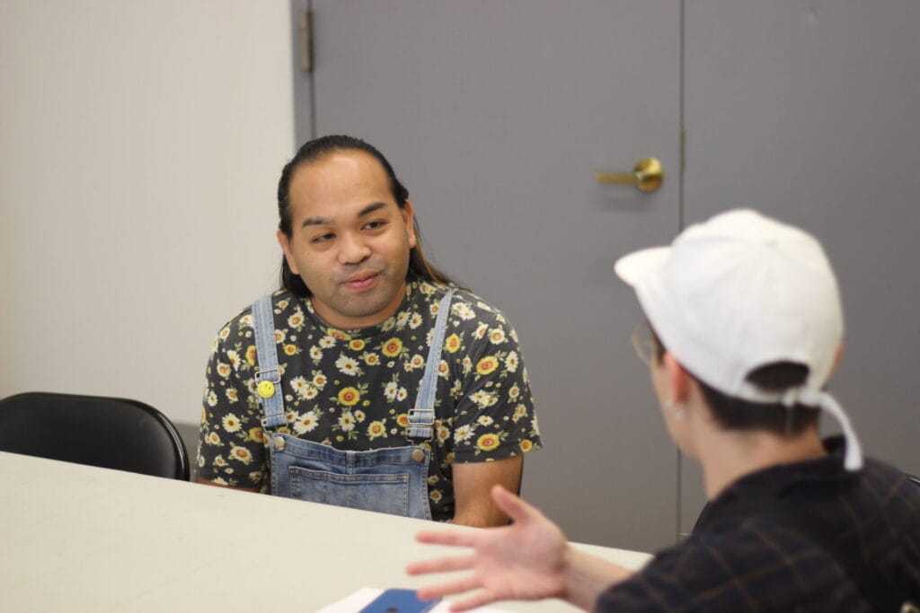 IMG_1231 | GLAAD Lana Leonard, white hat, dark flannel, interviews Edrian Pangilinan, long dark hair tied back, brown skin, with a yellow patterned shirt, adn overalls. The room is cold, white with grey doors.
